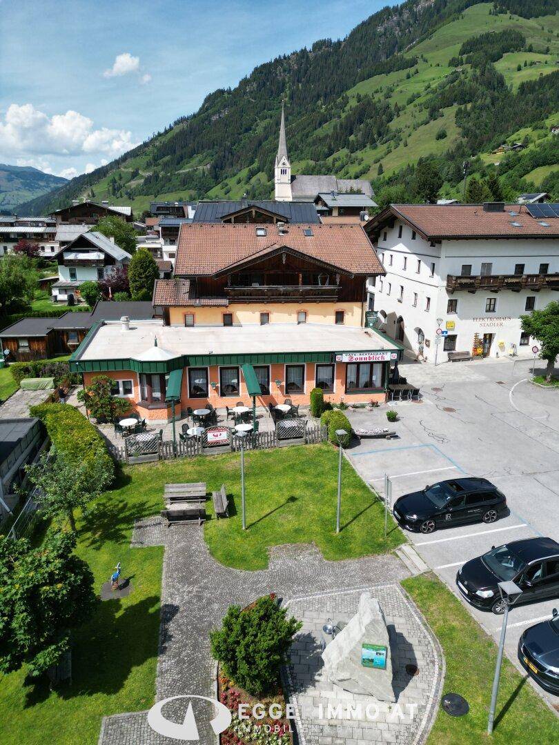 Panoramablick auf das alpine Dorf mit dem Restaurantgebäude, Parkplätzen und grünen Bergen.