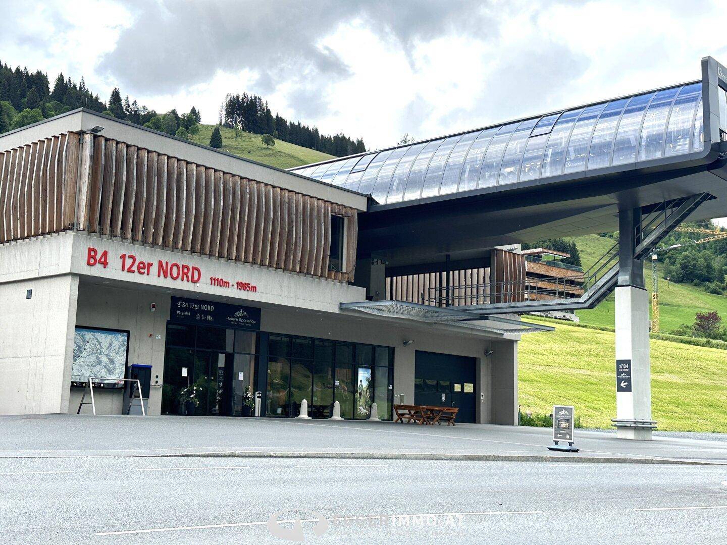 Modernes Gebäude einer Seilbahnstation mit Glas- und Holzelementen vor bergiger Kulisse.