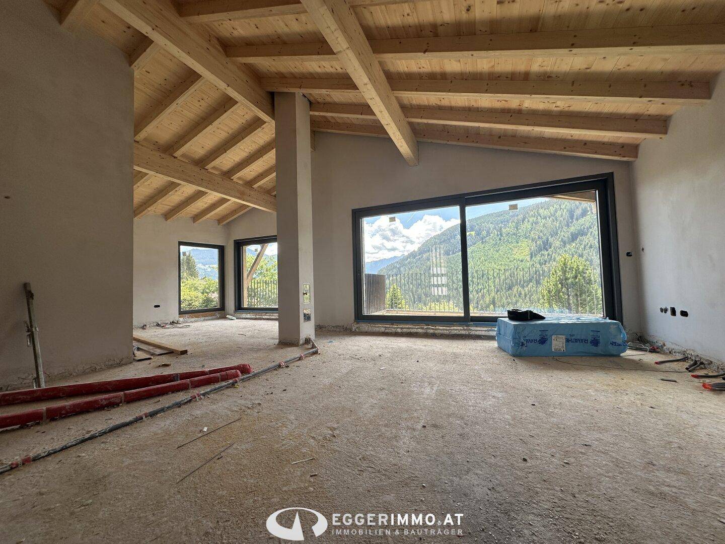 Heller Innenraum im Rohbau mit großen Fenstern und Bergblick, sichtbare Holzbalken an der Decke.