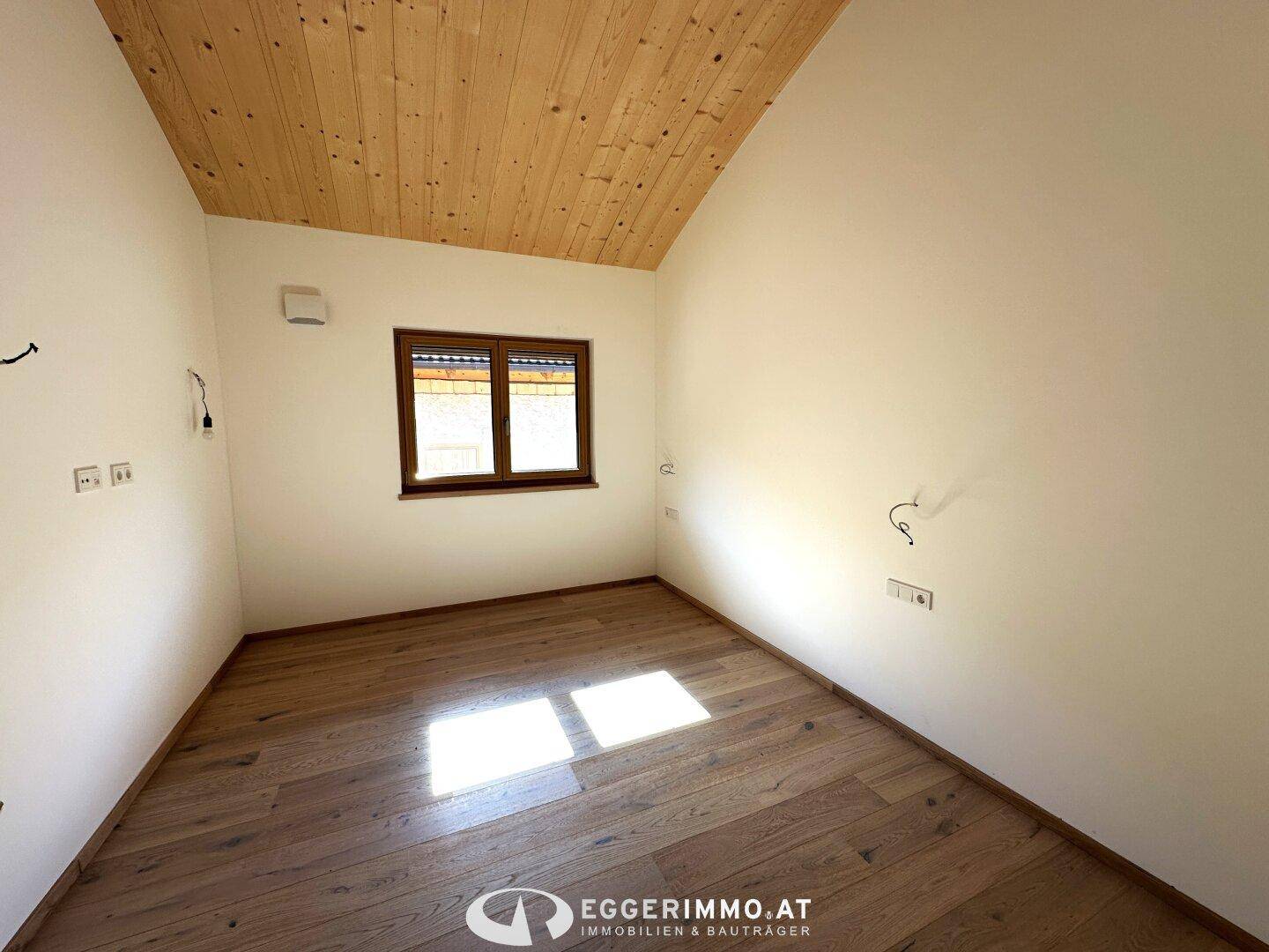 Helles Zimmer mit Holzboden, Holzdecke und einem Fenster, das viel Tageslicht hereinlässt.