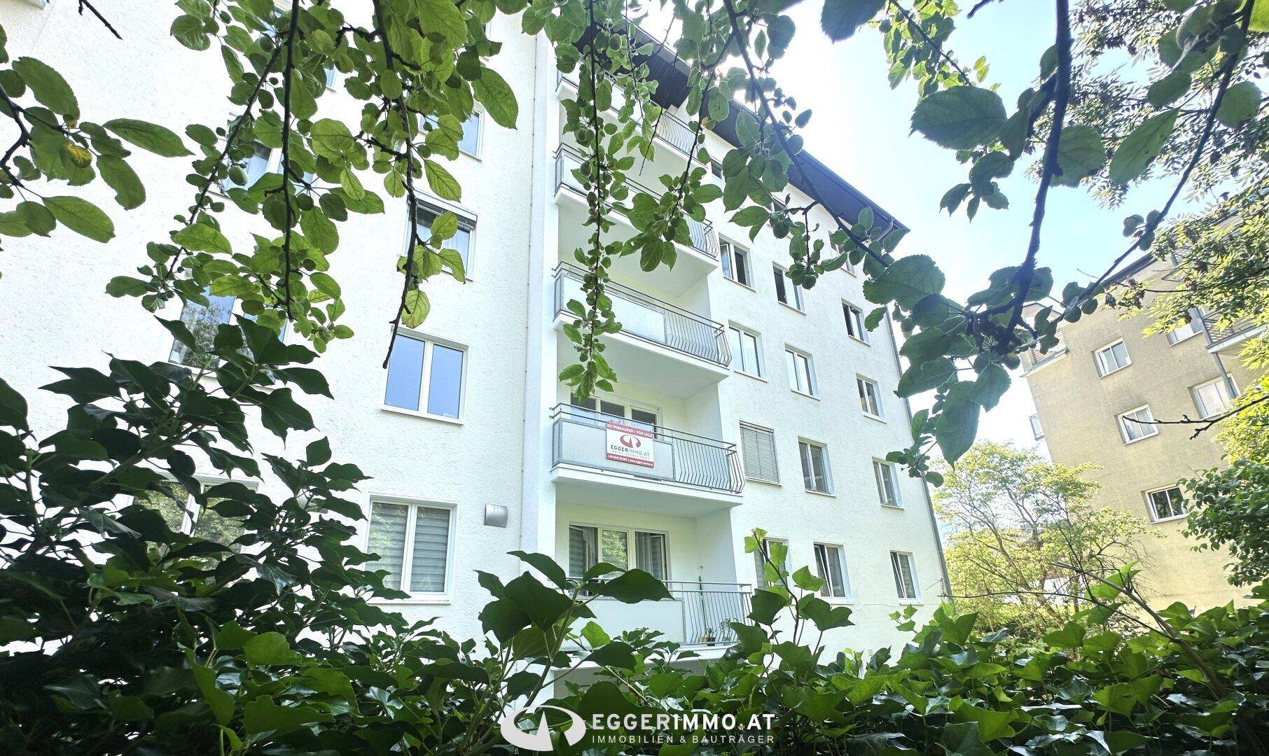 Wohngebäude mit Balkonen, teilweise verdeckt durch grüne Blätter, an einem sonnigen Tag.