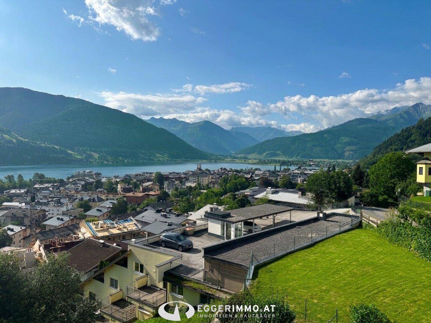 Weitläufiger Blick auf den See, die Stadt und die umliegende Berglandschaft mit grünen Wiesen.