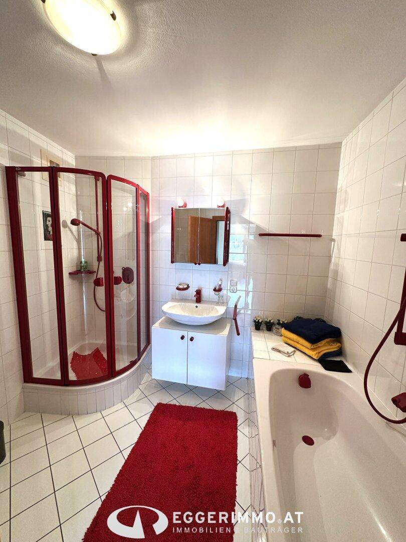 Badezimmer mit Badewanne, Duschkabine und weißen Fliesen, akzentuiert mit roten Details.