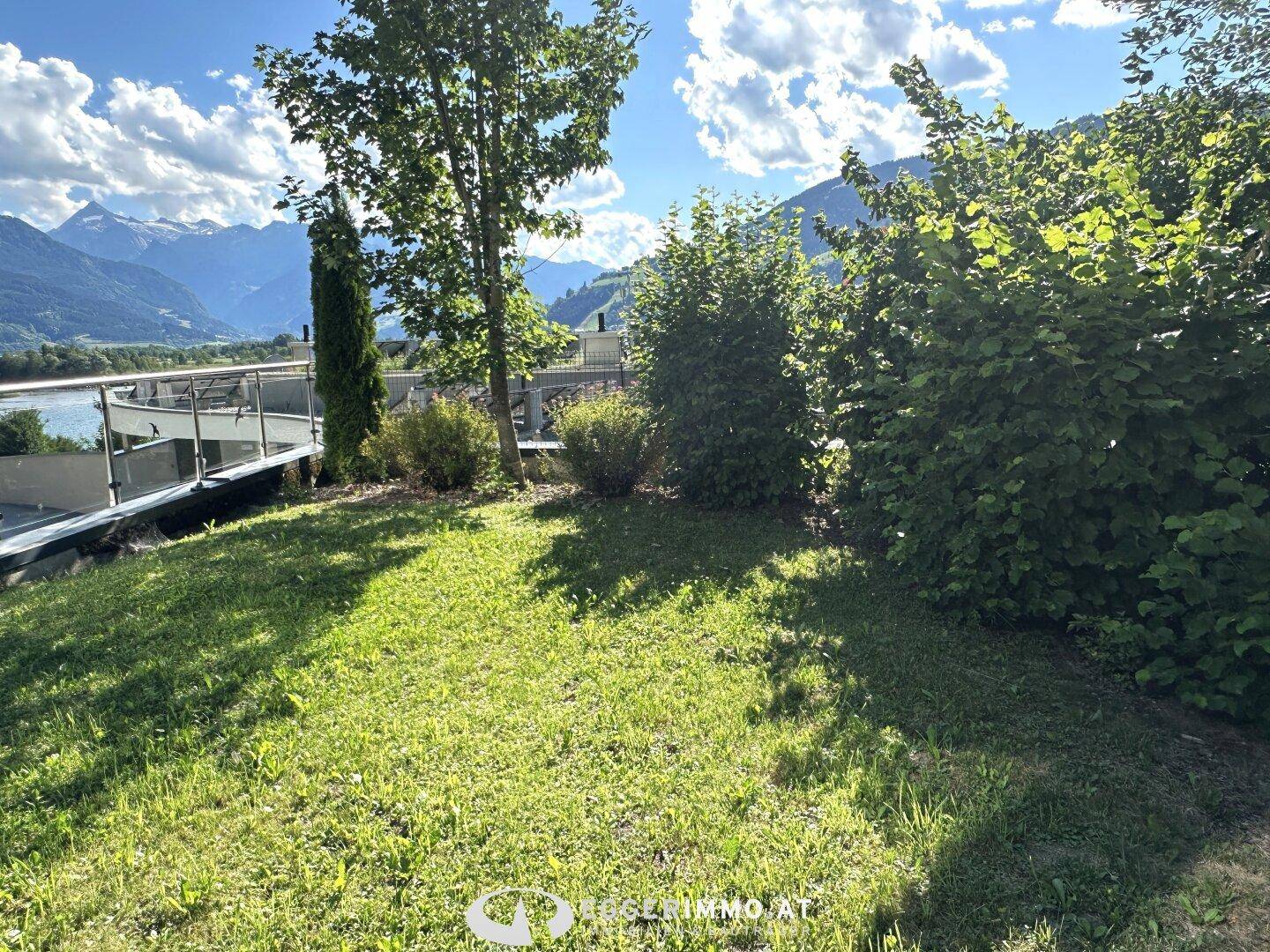 Grüne Gartenfläche mit üppigen Büschen und Bäumen, die einen malerischen Blick auf den See und die Berge freigibt.