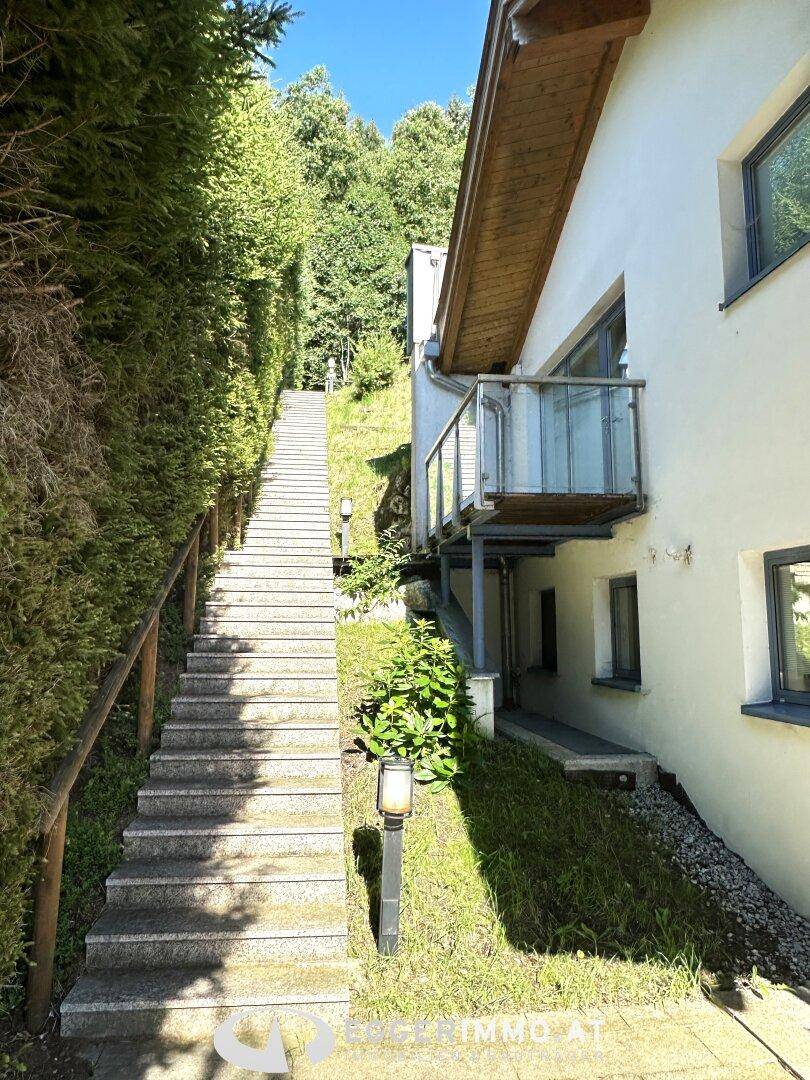Lange Steintreppe führt einen bewaldeten Hang hinauf, flankiert von einer Hausfassade mit einem kleinen Balkon.