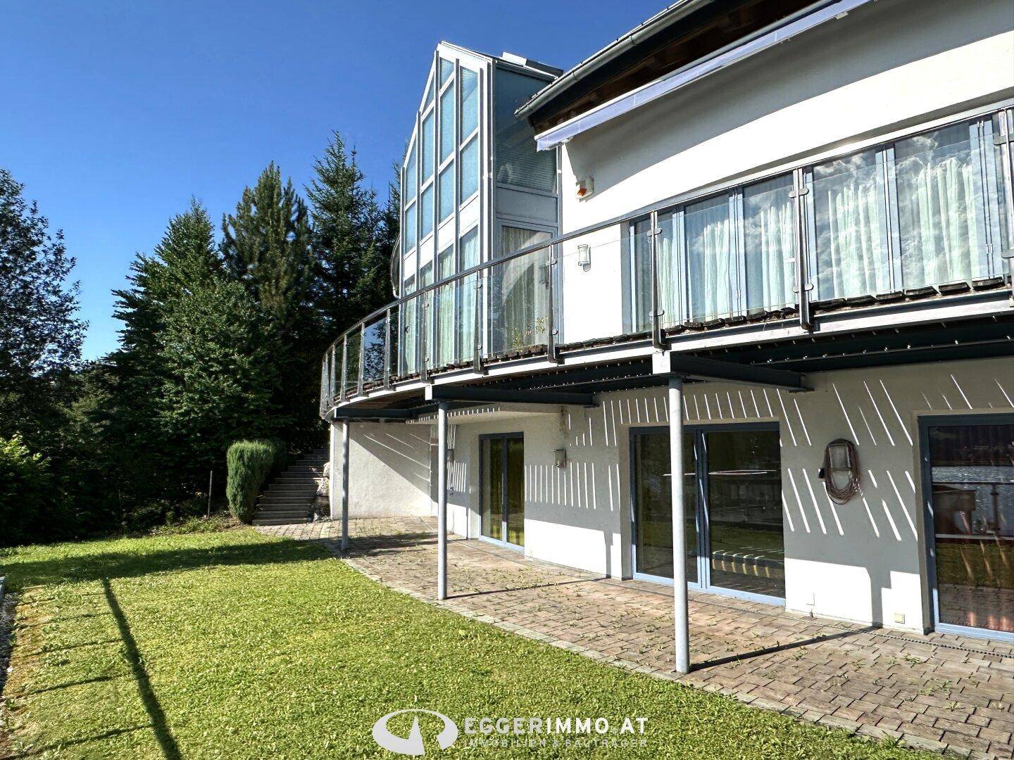 Moderne Hausfassade mit großen Glasflächen und einem geschwungenen Balkon über einer gepflegten Rasenfläche.