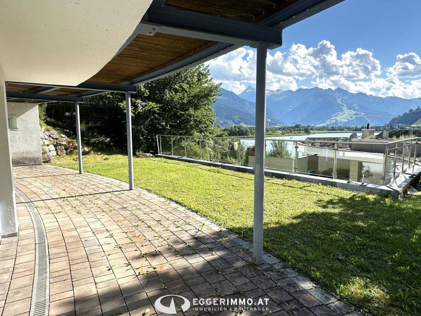 Große gepflasterte Terrasse mit Rasenfläche und Glasgeländer, die einen weiten Blick auf Berge und See bietet.