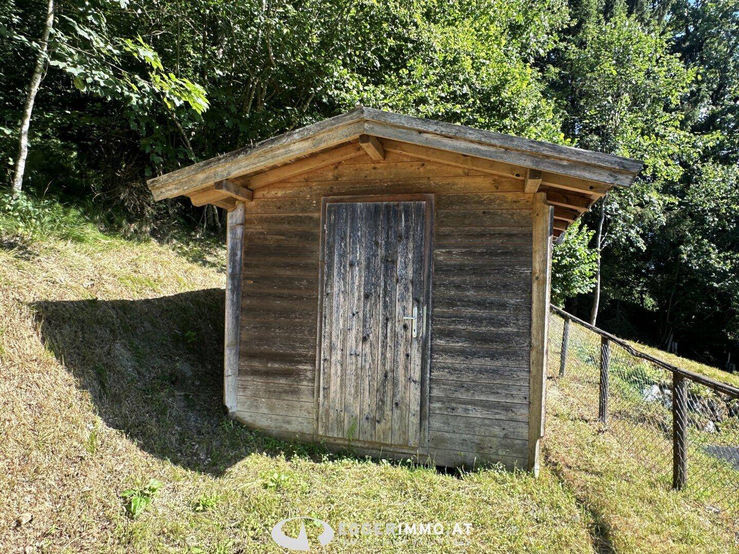 Rustikaler hölzerner Gartenschuppen auf einer sonnigen Wiese, umgeben von Bäumen und Sträuchern.