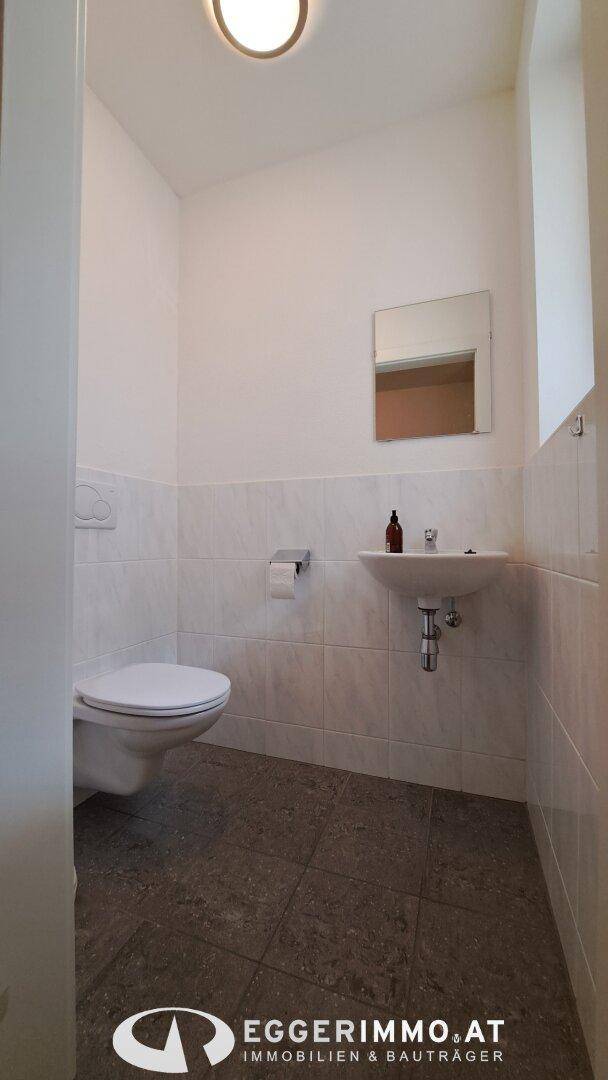 Gäste-WC mit Wand-WC, Waschbecken und Spiegel, halbhoch gefliest.