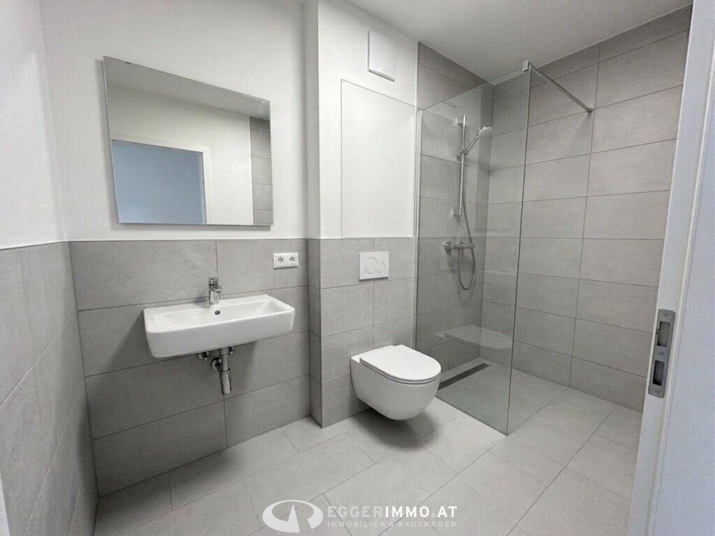 Modernes Badezimmer mit grauen Fliesen, wandhängendem WC und begehbarer Dusche.