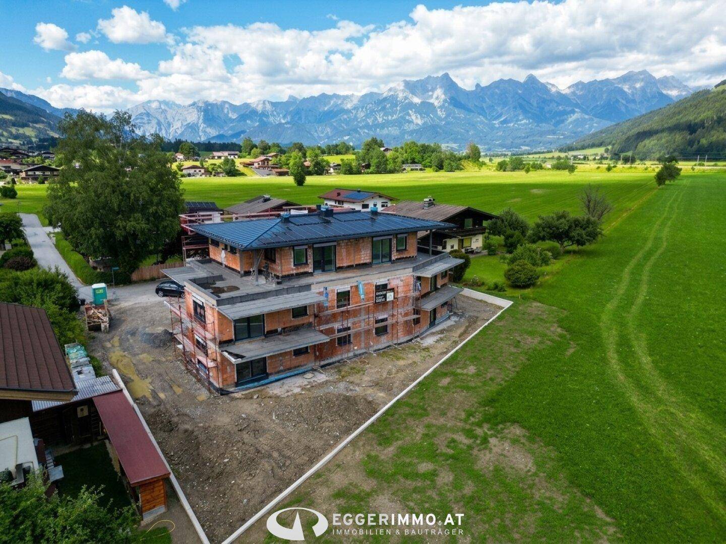 Luftaufnahme eines im Bau befindlichen Gebäudes inmitten grüner Felder mit majestätischem Bergpanorama.