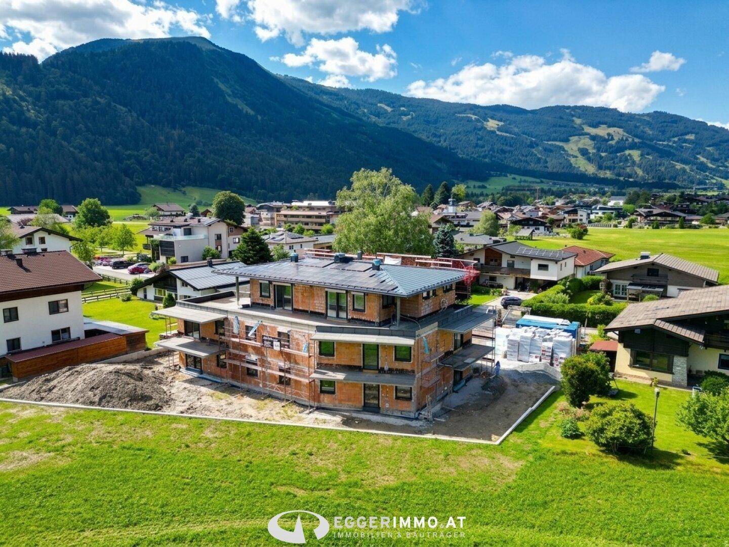 Luftaufnahme eines Neubaus im Rohbau, umgeben von grünen Wiesen und Bergen.