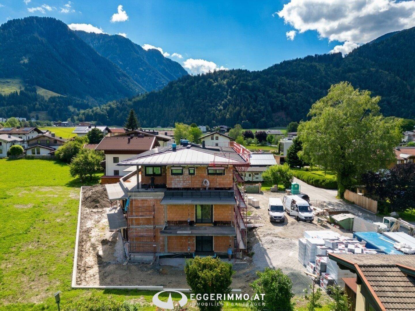 Luftaufnahme eines im Bau befindlichen Gebäudes mit Baustellenfahrzeugen und Bergkulisse.
