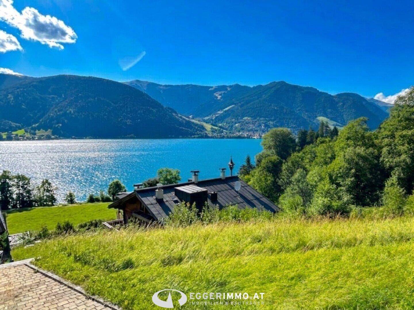 Idyllischer Blick auf ein Haus mit dunklem Dach, umgeben von grünen Wiesen, mit einem See und Bergen im Hintergrund.
