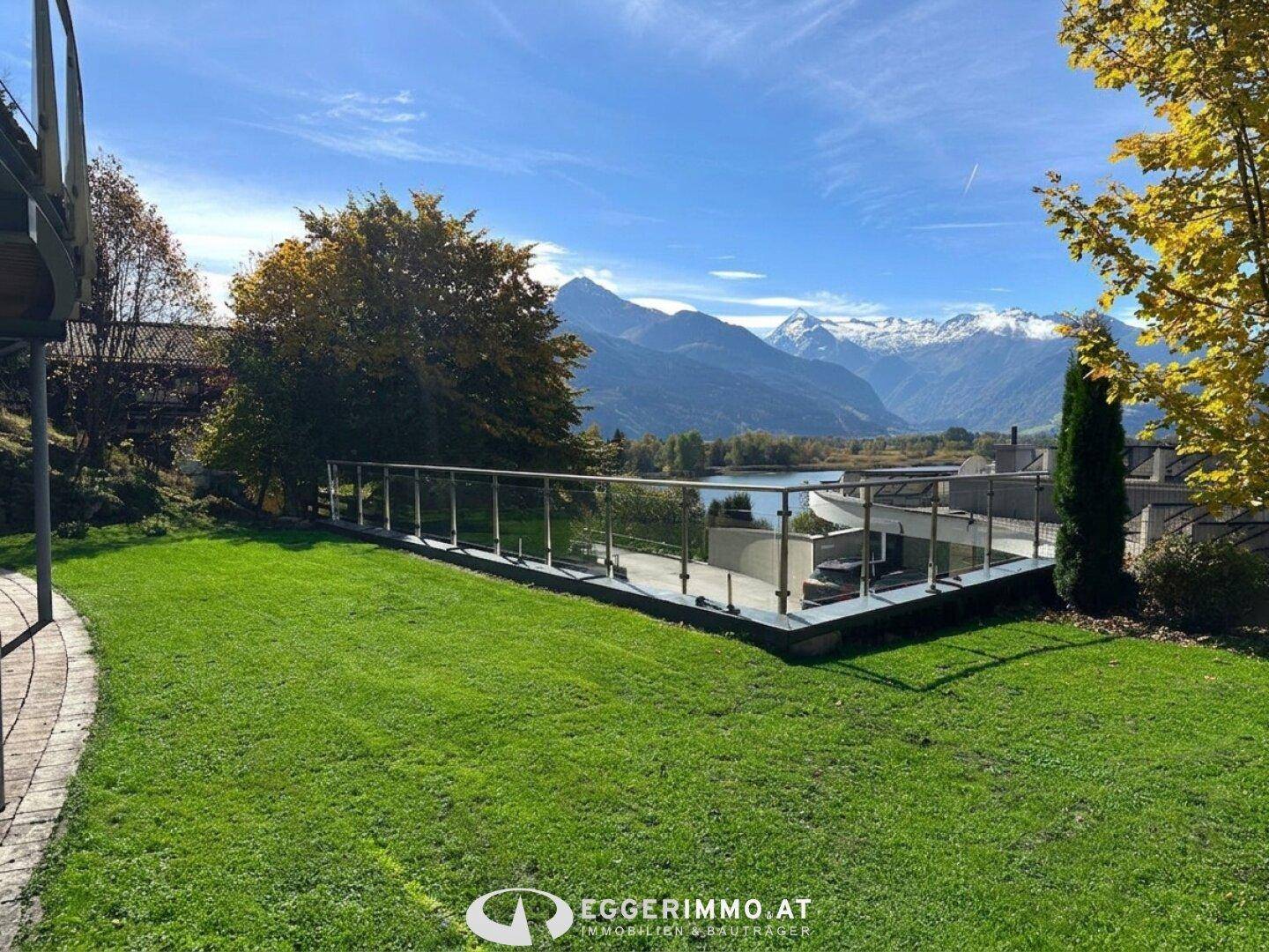 Gepflegter Gartenbereich mit Rasen und einer modernen Terrasse mit Glasgeländer, Blick auf See und Berge.