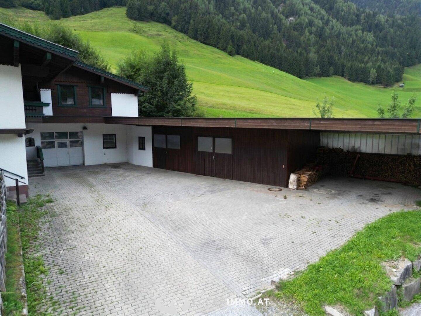 Gepflasterter Bereich neben einem Gebäude mit Holzlager und einer Zufahrt.