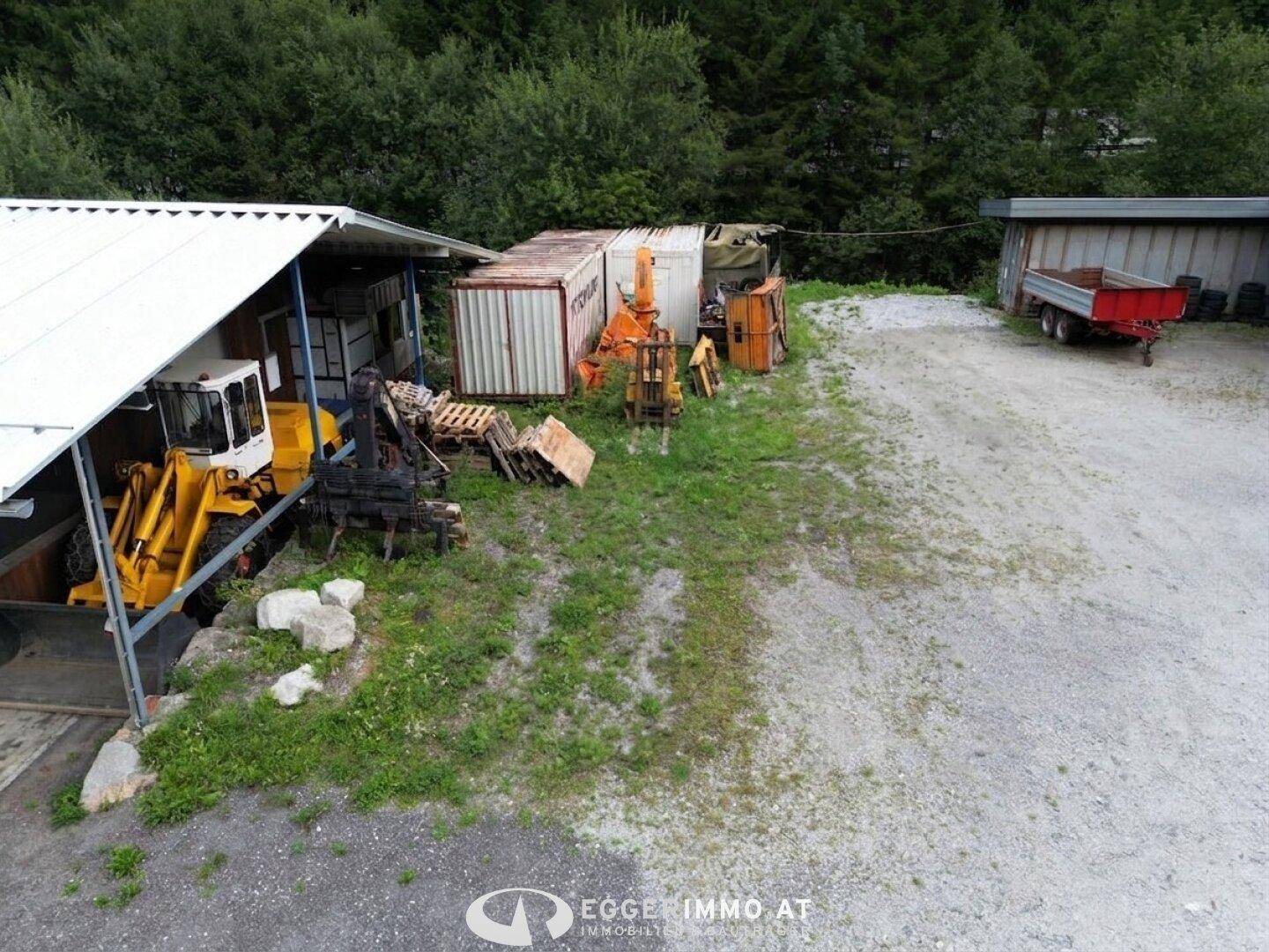 Außenbereich eines Gewerbebetriebs mit Baumaschinen, Lagercontainern und Schotterfläche.