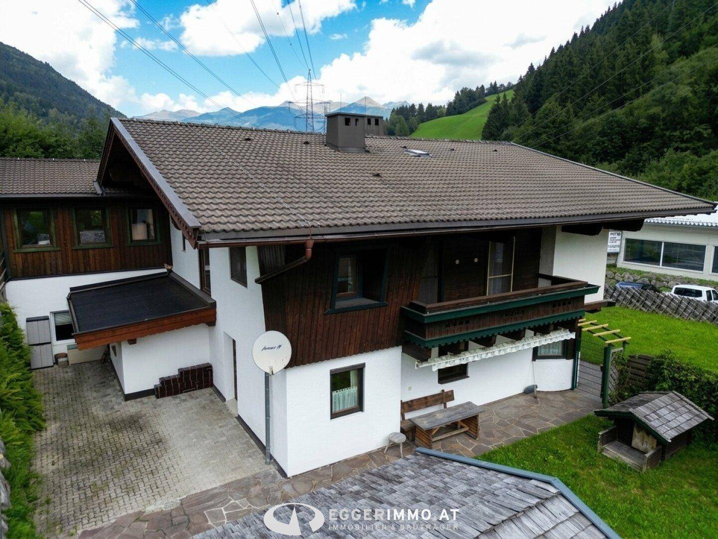 Traditionelles Wohnhaus mit Balkon, Satellitenschüssel und gepflastertem Vorplatz vor grüner Kulisse.