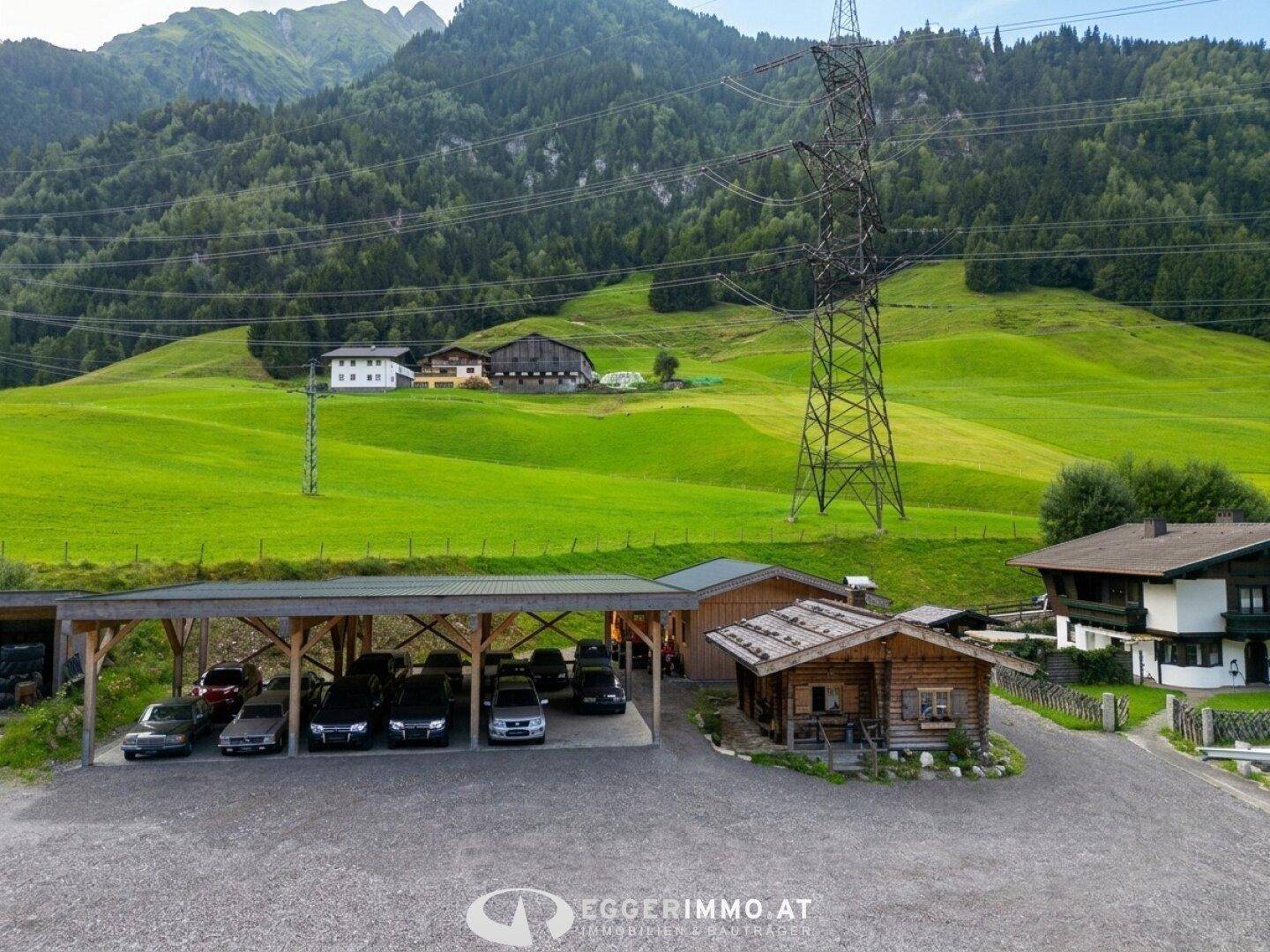Weitläufiges Anwesen mit Carport, Holzhütte und traditionellem Haus inmitten grüner Wiesen und Berge.