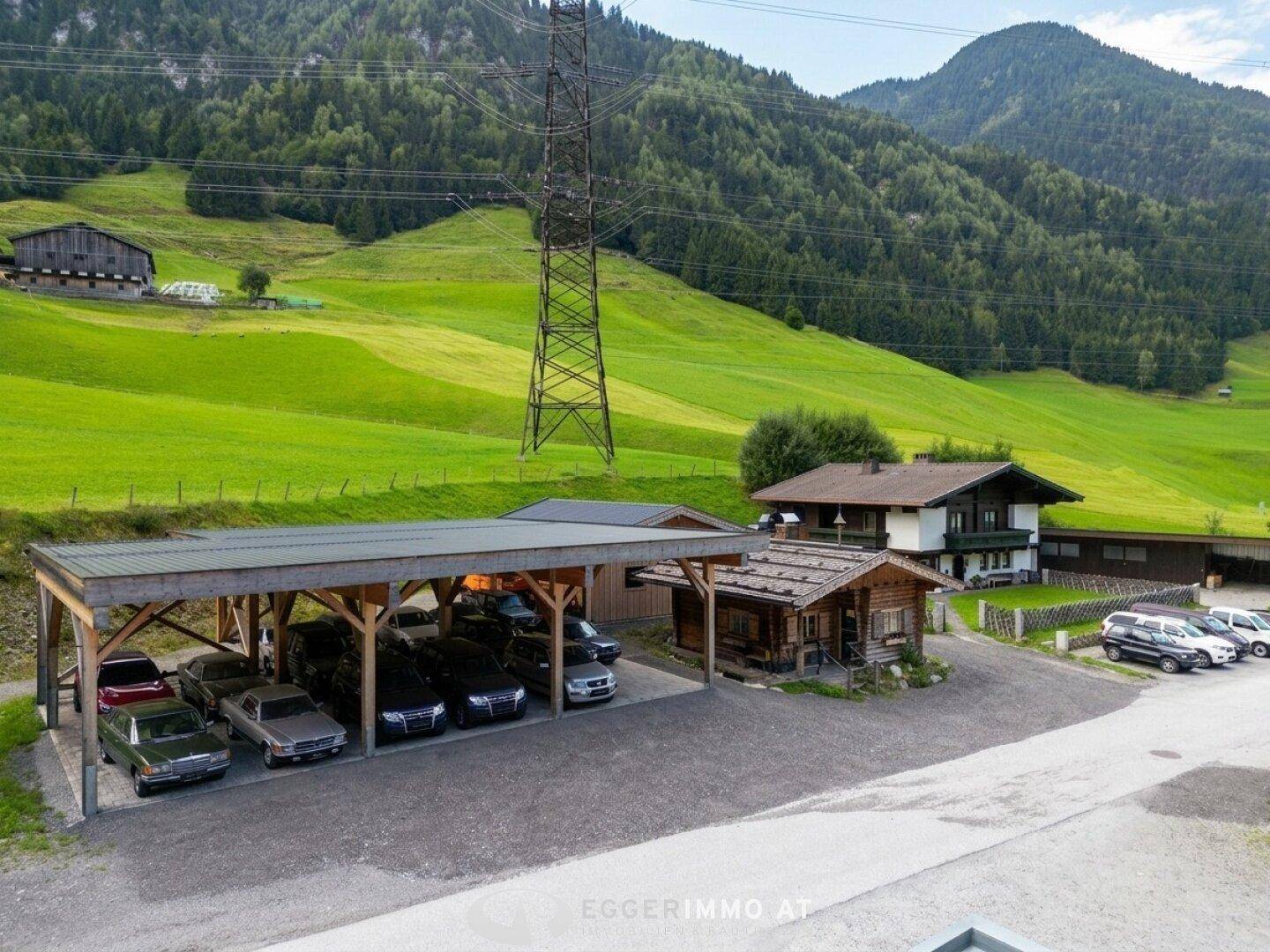 Panoramablick auf ein ländliches Anwesen mit Carport, Holzhütte und Wohnhaus, umgeben von grünen Hügeln.