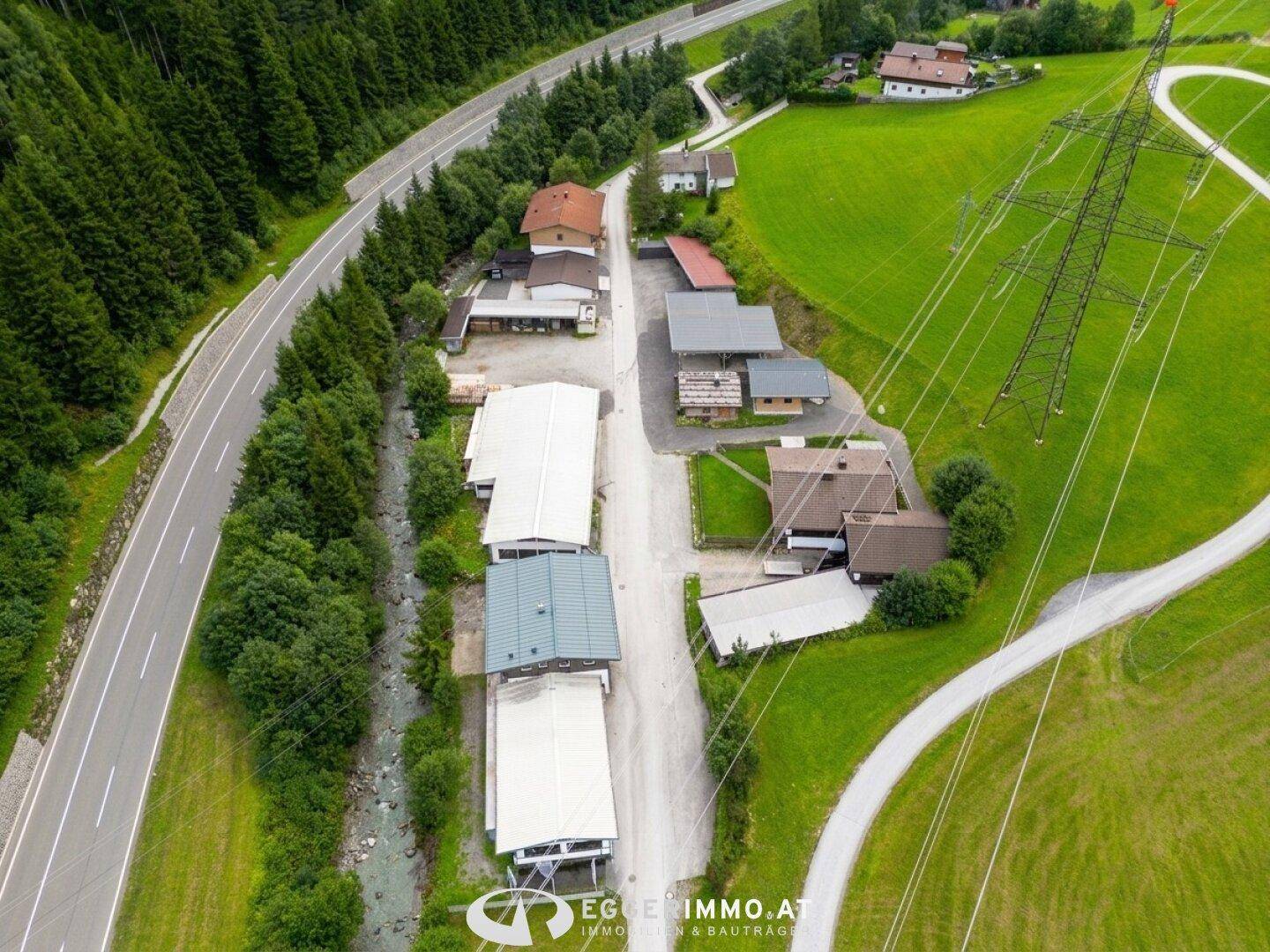 Weitläufige Luftaufnahme des Anwesens mit angrenzender Straße, Fluss und grüner Berglandschaft.