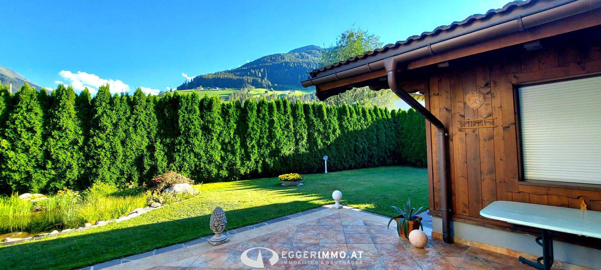 Sonniger Gartenbereich mit Terrasse, Teich und hoher Hecke vor Bergkulisse.