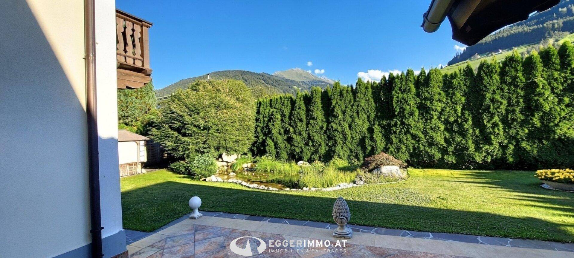 Gepflegter Garten mit Teich, Rasenfläche und einer Terrasse mit Bergblick.