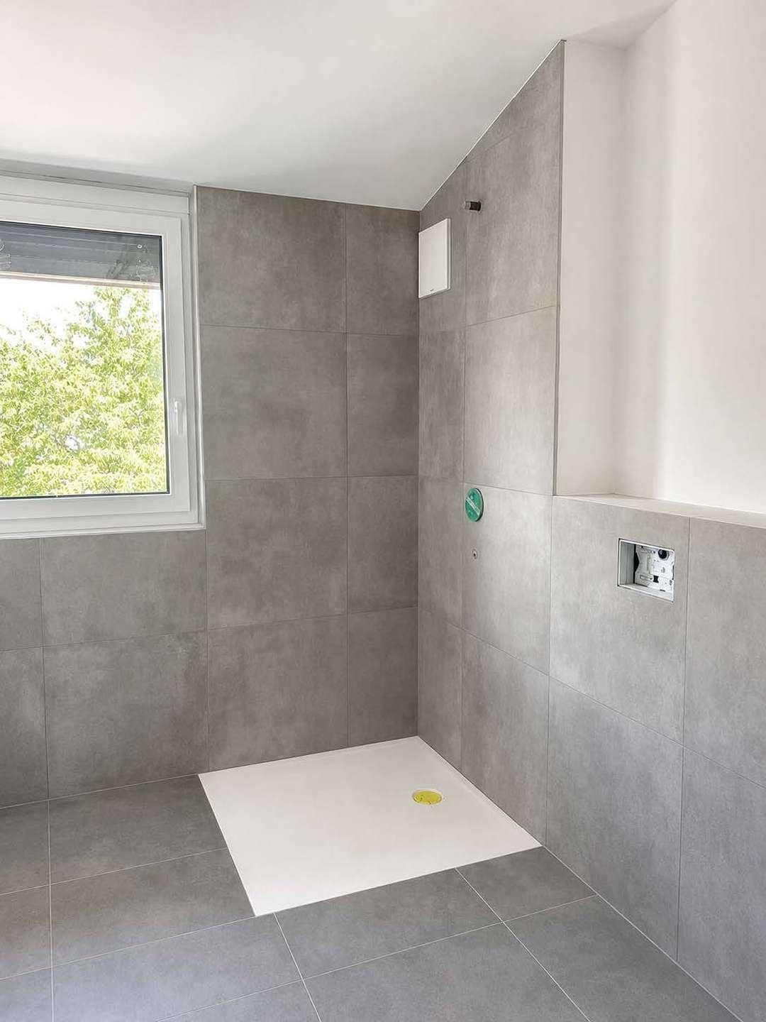 Modernes Badezimmer im Bau mit grauen Fliesen, Duschbereich und Fenster.