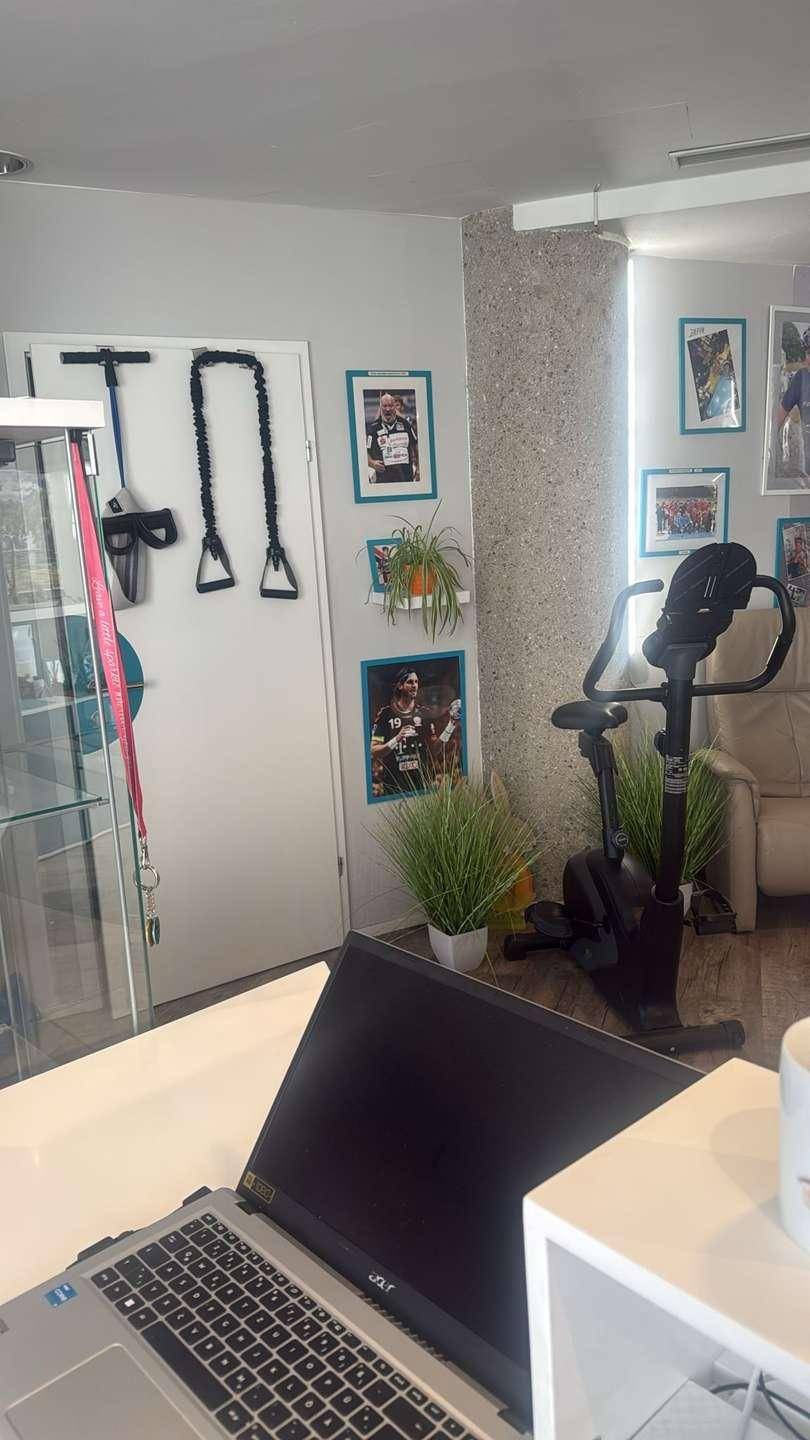 Trainingsbereich mit Heimtrainer und Laptop auf einem Schreibtisch, dekoriert mit gerahmten Bildern.