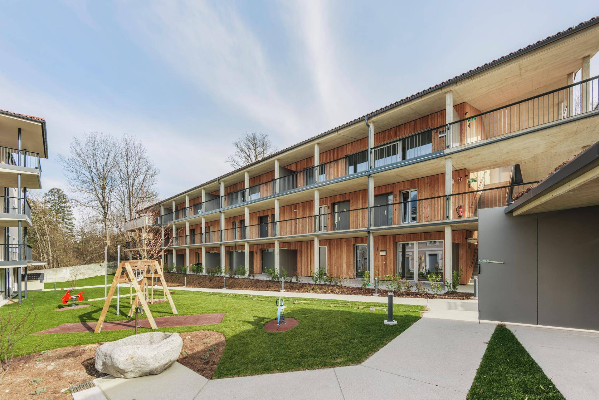 Zeitgenössisches Wohngebäude mit Holzverkleidung, großzügigen Balkonen und einem Gemeinschaftsspielplatz.