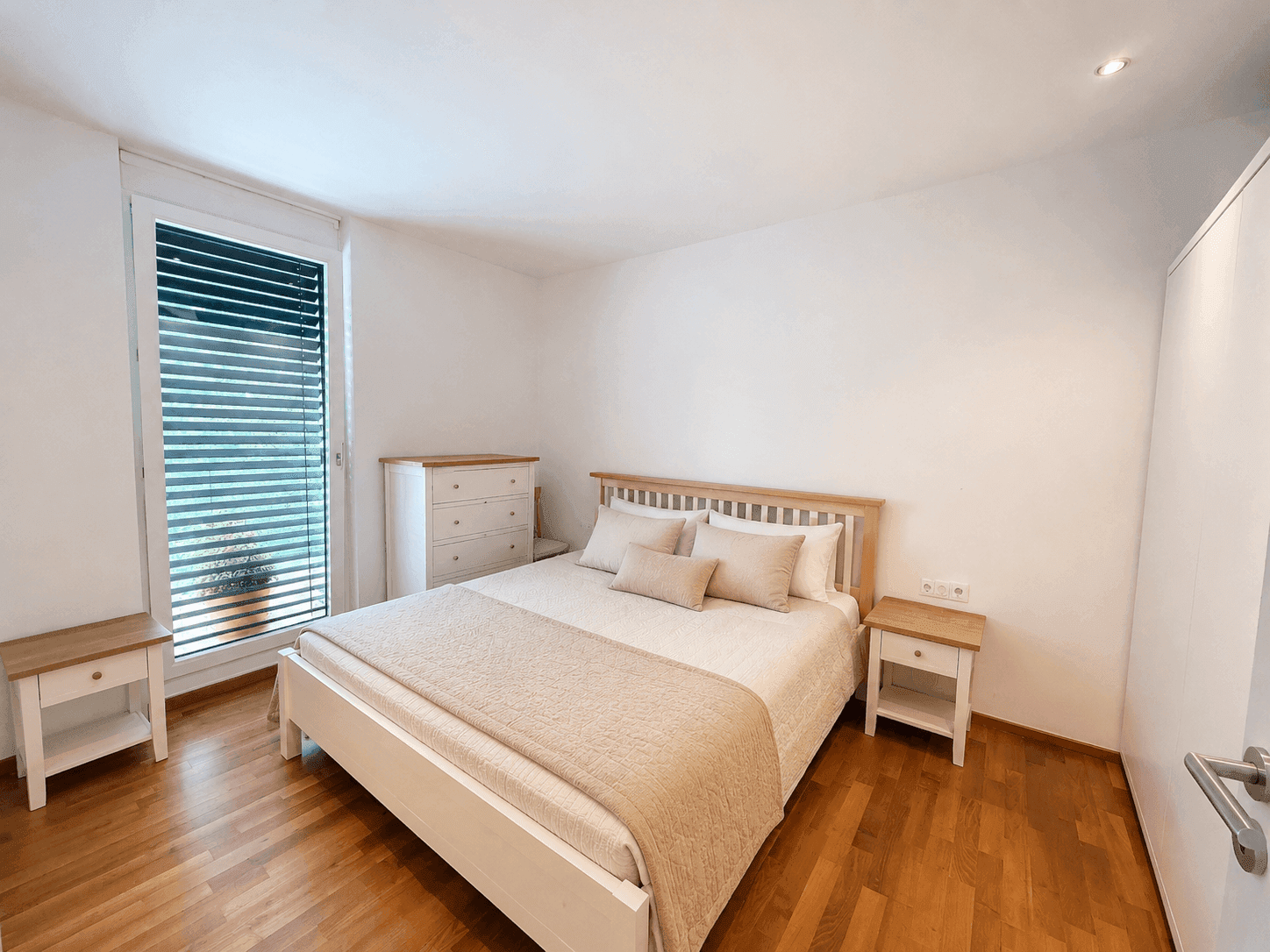 Helles Schlafzimmer mit Holzboden, Doppelbett und großem Fenster mit Jalousien.