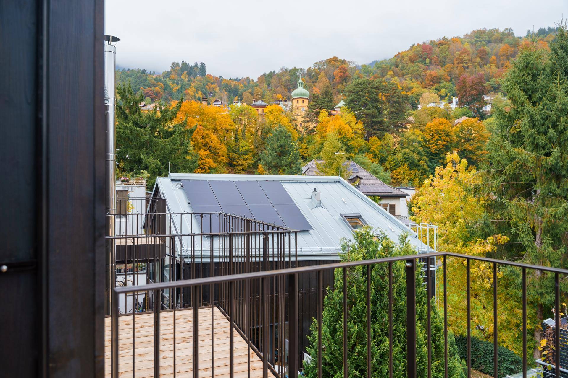 Balkon mit Holzbelag und Blick auf umliegende Gebäude, herbstliche Bäume und einen Kirchturm.
