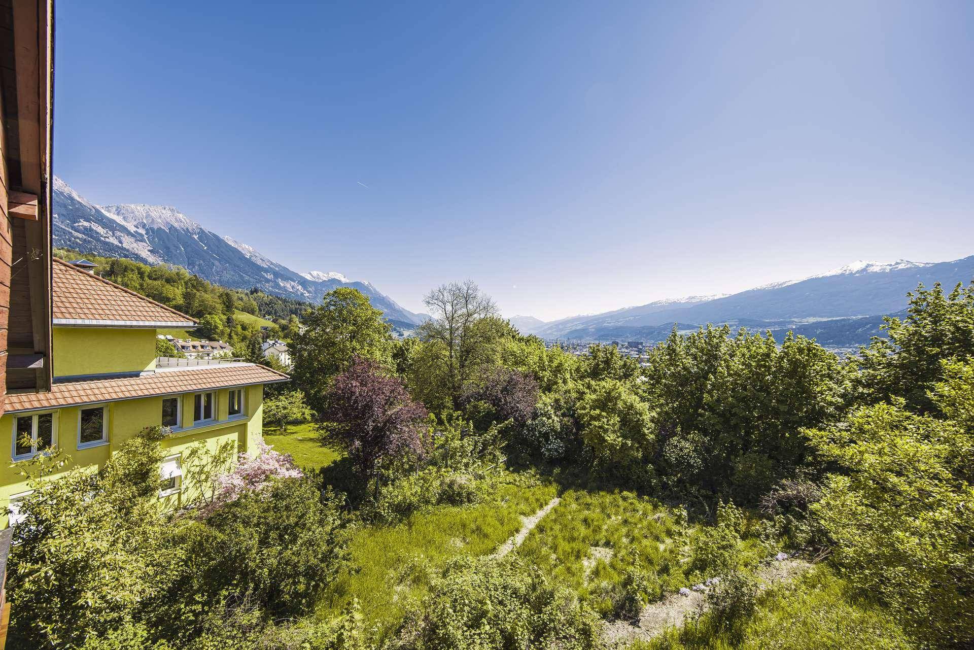 Weitläufiger Ausblick auf die grüne Landschaft, ein Nachbargebäude und die beeindruckende Bergkulisse.