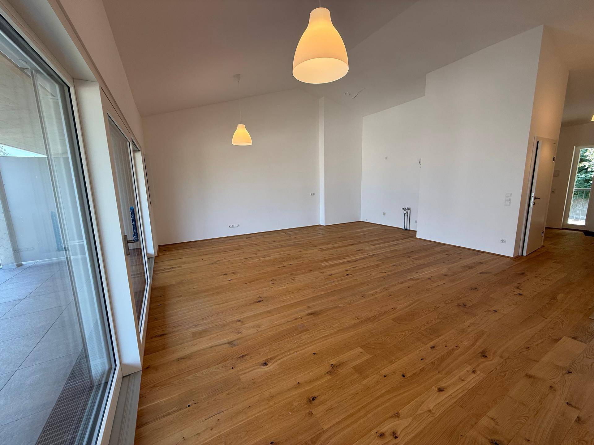 Geräumiger Wohnbereich mit Holzboden und großen Fenstern, die auf einen Balkon führen.