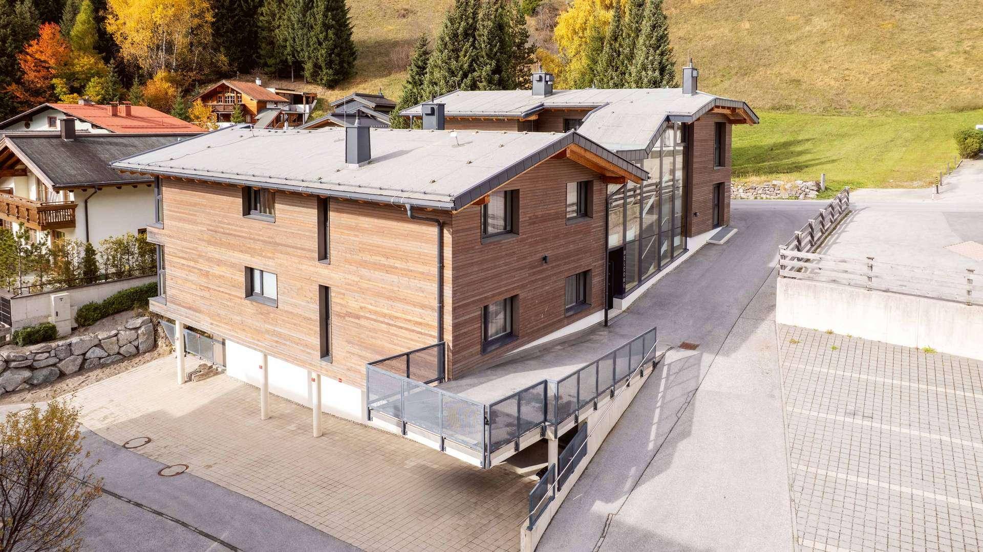 Gebäudeansicht von oben mit Holzfassade, Glasfront und Parkplätzen in den Bergen.
