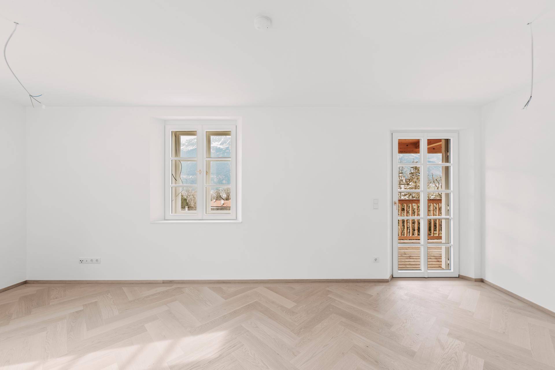 Lichtdurchfluteter Raum mit Fischgrätparkett, großem Fenster und Zugang zu einem Außenbereich.