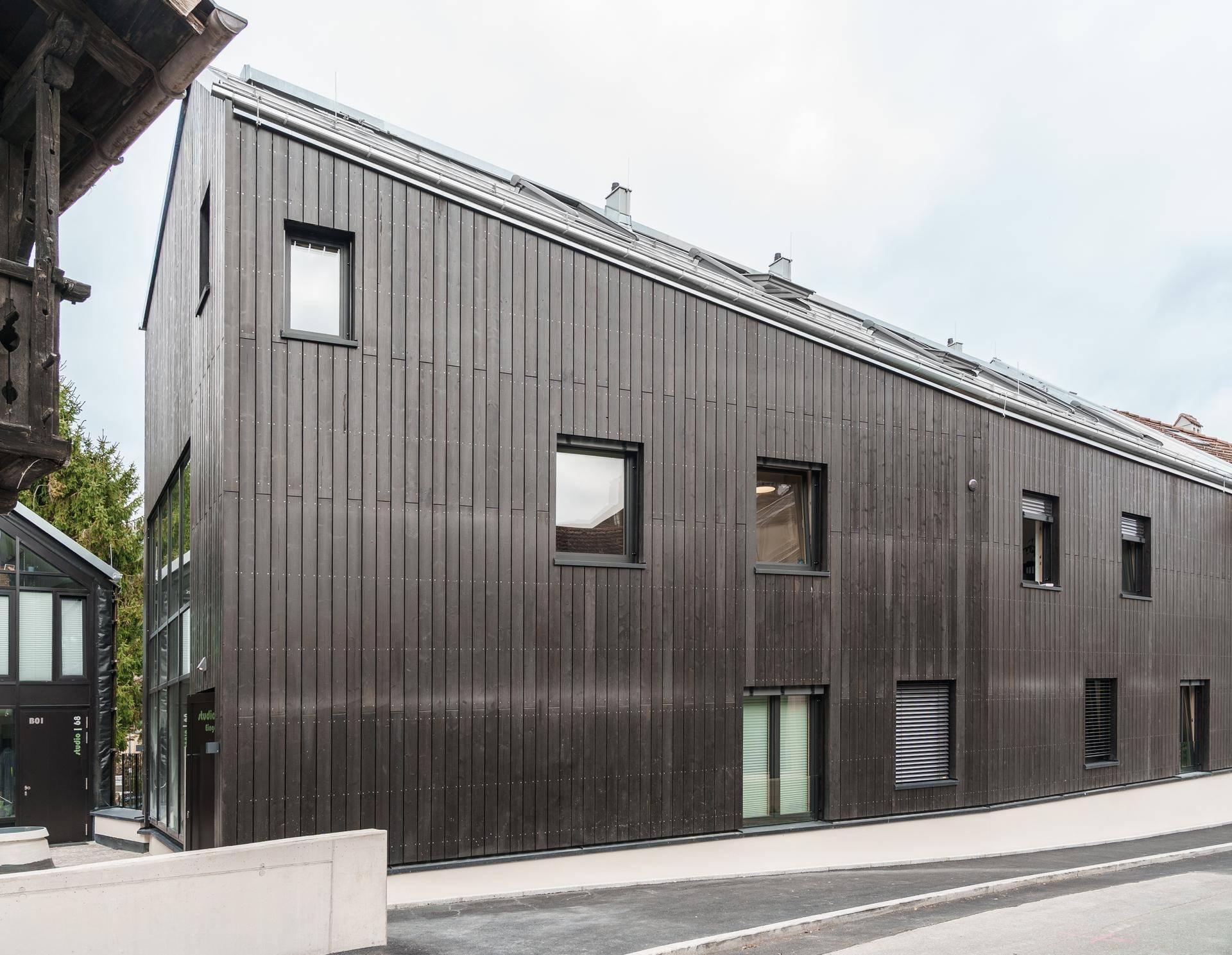 Schräge Ansicht des modernen Gebäudes mit dunkler Holzfassade und schräger Dachlinie neben einem Altbau.