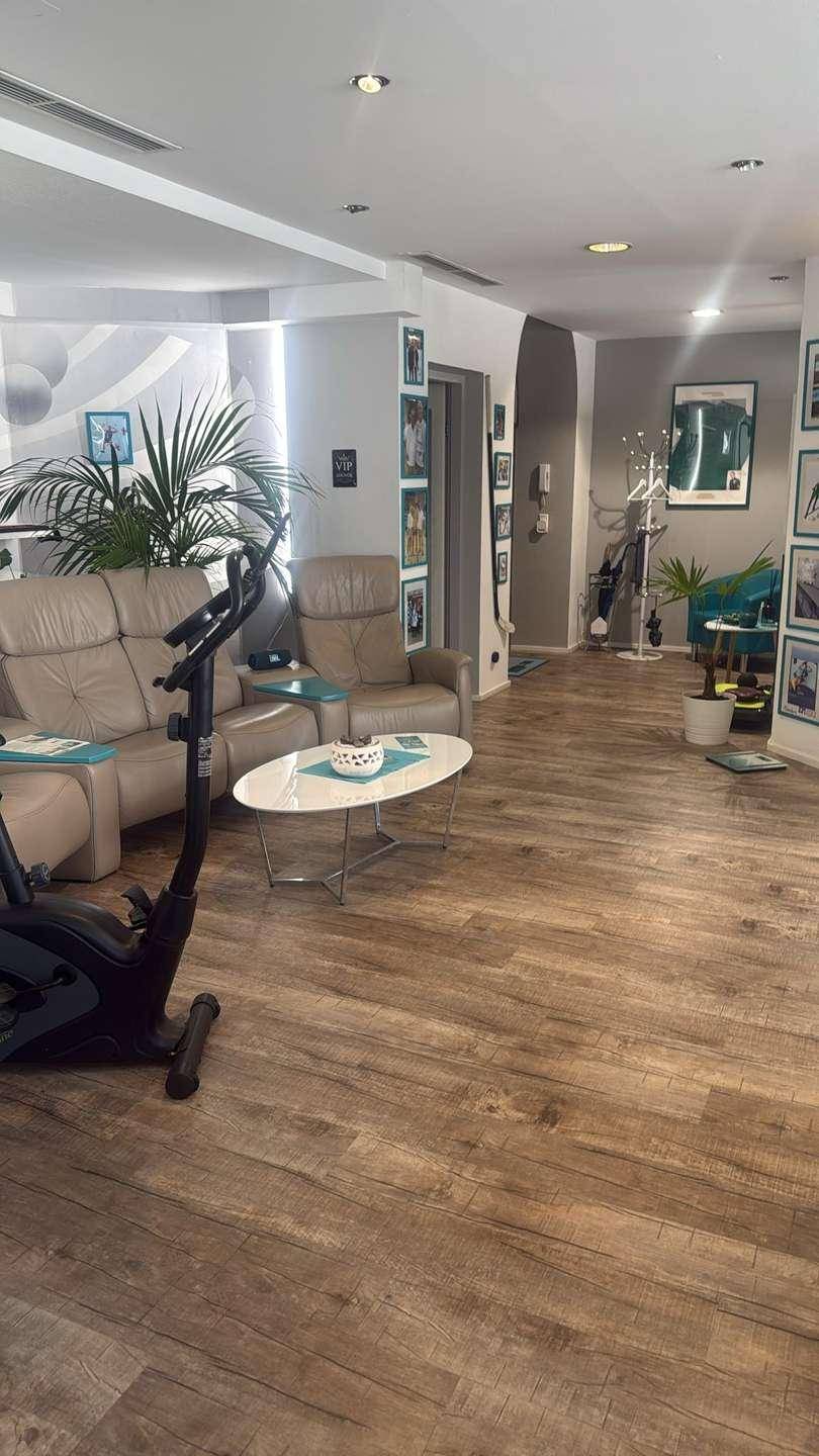 Wartebereich mit bequemen Sesseln, Couchtisch und Fitnessgeräten im Vordergrund, hell und geräumig.