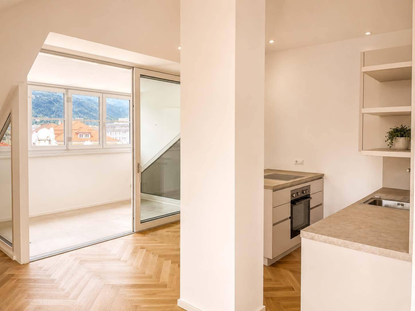 Heller Wohnbereich mit Fischgrätparkett, Blick auf die Berge und Zugang zu einem verglasten Balkon.