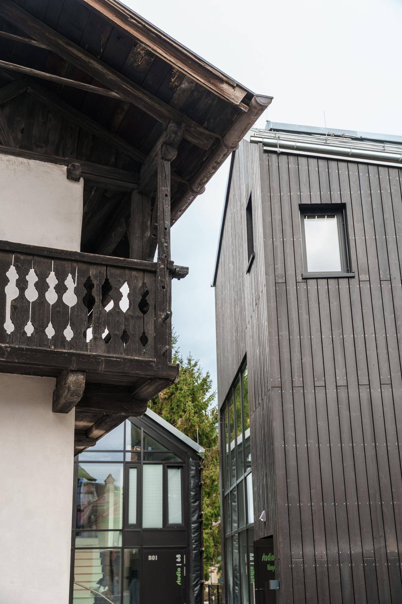 Kontrast zwischen einem traditionellen Gebäude mit Holzbalkon und einem modernen Neubau mit dunkler Holzfassade.