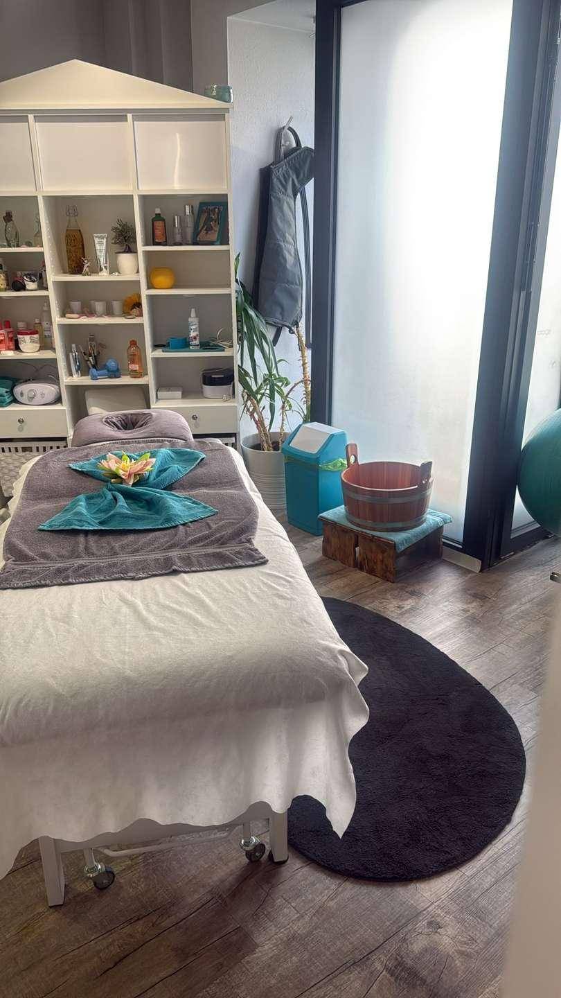 Behandlungsraum mit Massageliege, weißem Regal und großem Fenster, das viel natürliches Licht hereinlässt.
