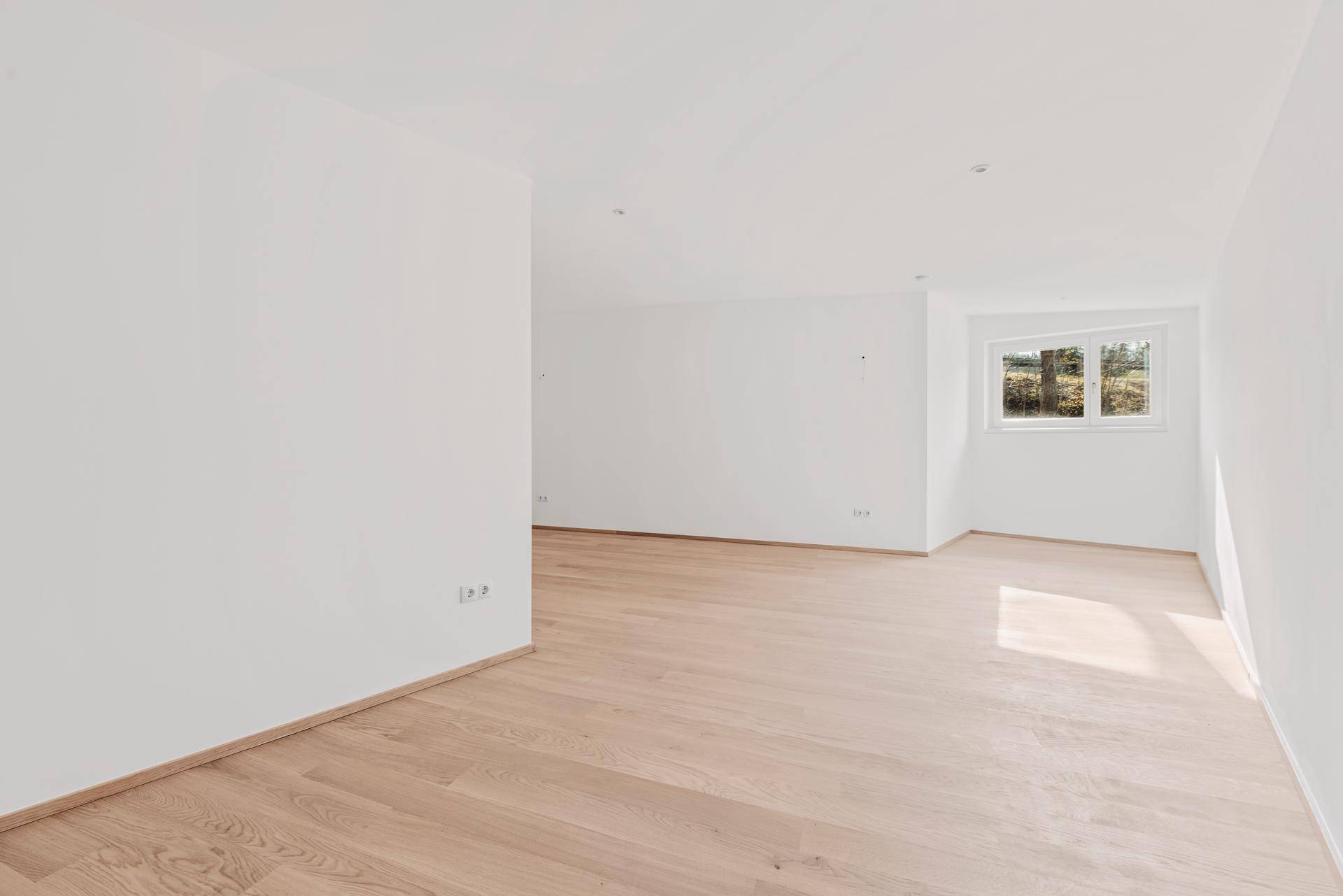 Leerer Raum mit Parkettboden und einem Fenster, das natürliches Licht hereinlässt.