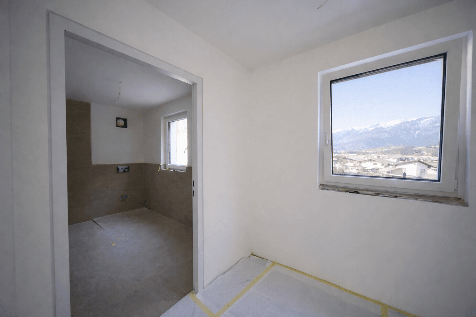 Unfertiger Raum mit Fenster und Bergblick, angrenzend an ein Badezimmer im Rohbau.