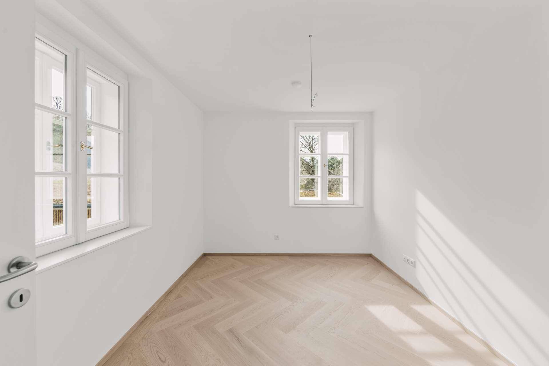 Leeres Schlafzimmer mit Fischgrätparkett und zwei großen Fenstern.