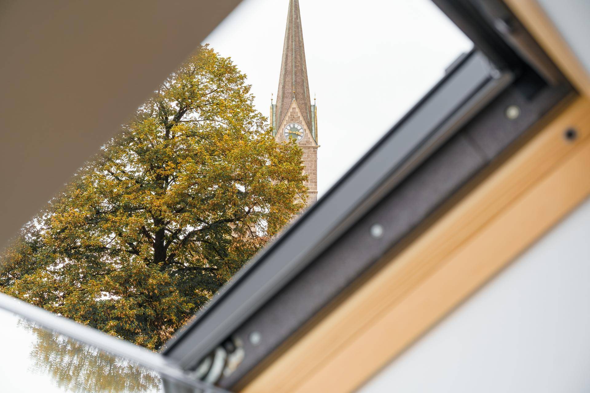 Blick durch ein Dachfenster auf einen Kirchturm und bunte Herbstbäume.
