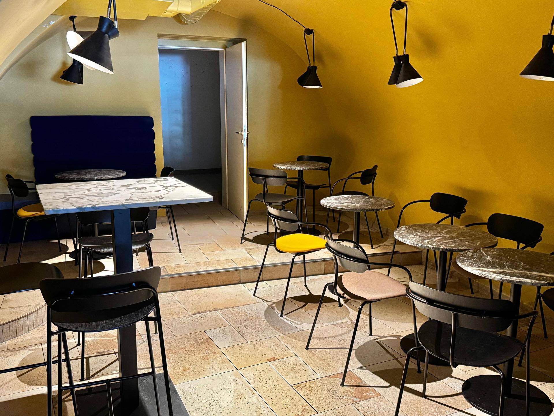 Gemütlicher Sitzbereich mit gelben Wänden, verschiedenen Tischen und modernen Hängelampen in einem Café.