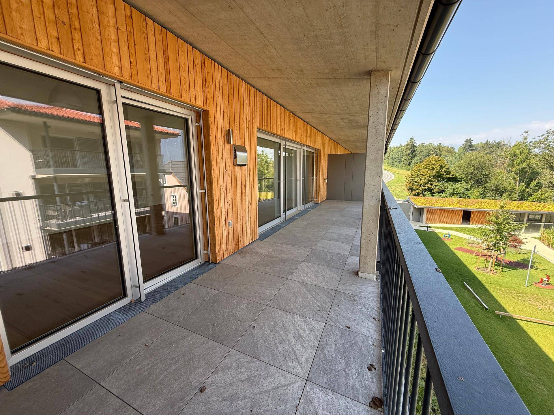 Großer Balkon mit Fliesenboden, Holzfassade und Blick auf eine grüne Fläche mit Spielplatz.