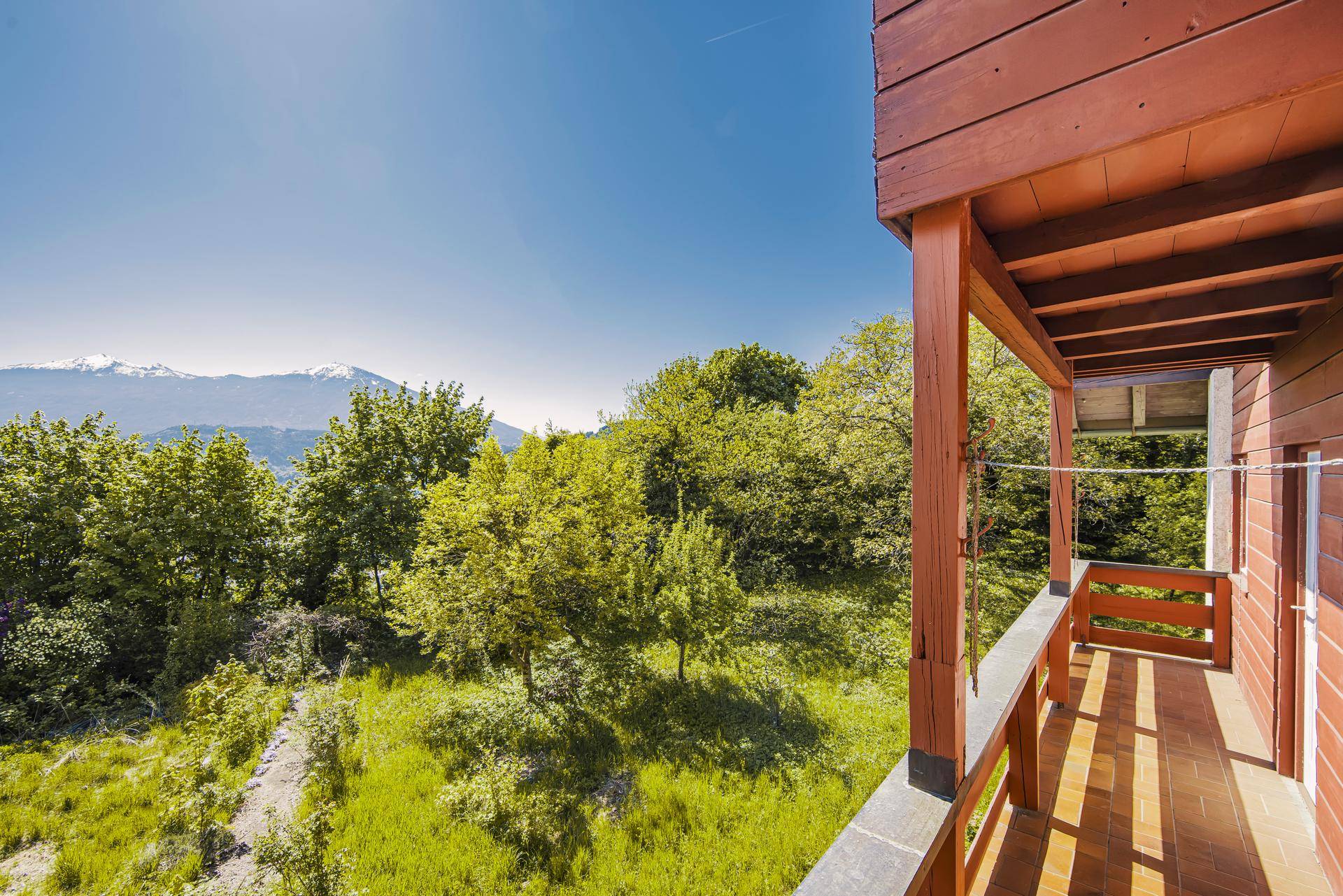 Hölzerner Balkon mit weitem Blick über grüne Bäume und schneebedeckte Berge unter blauem Himmel.