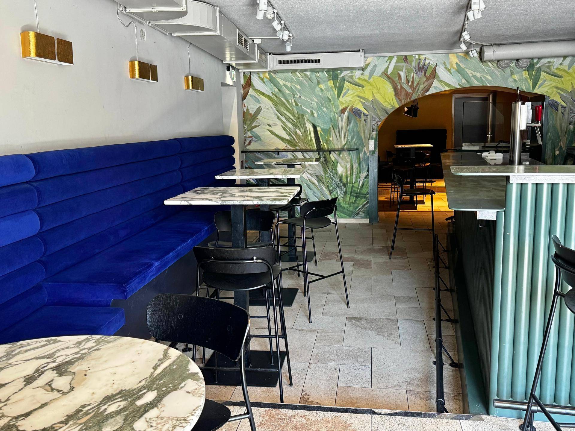 Stilvoller Innenraum einer Bar mit blauen Sitzgelegenheiten, Stehtischen und einer tropischen Wandmalerei.