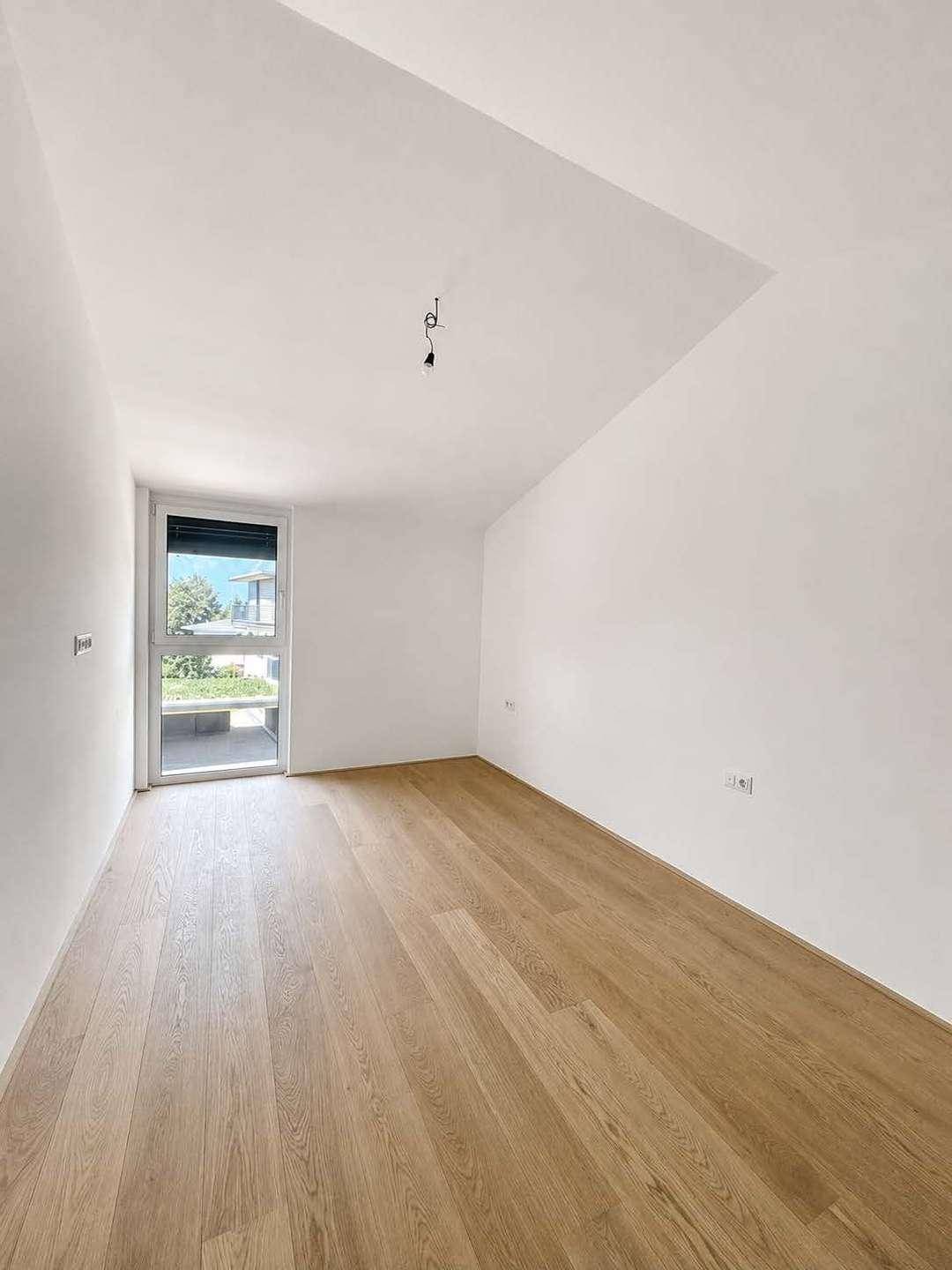 Helles Zimmer mit Holzboden und großem Fenster mit Zugang zum Außenbereich.